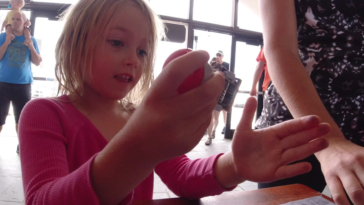In Winter Park, Colorado, the child holds the small camera close to her face as an adult hand opens beneath it and visitors stand near the glass entrance behind them.