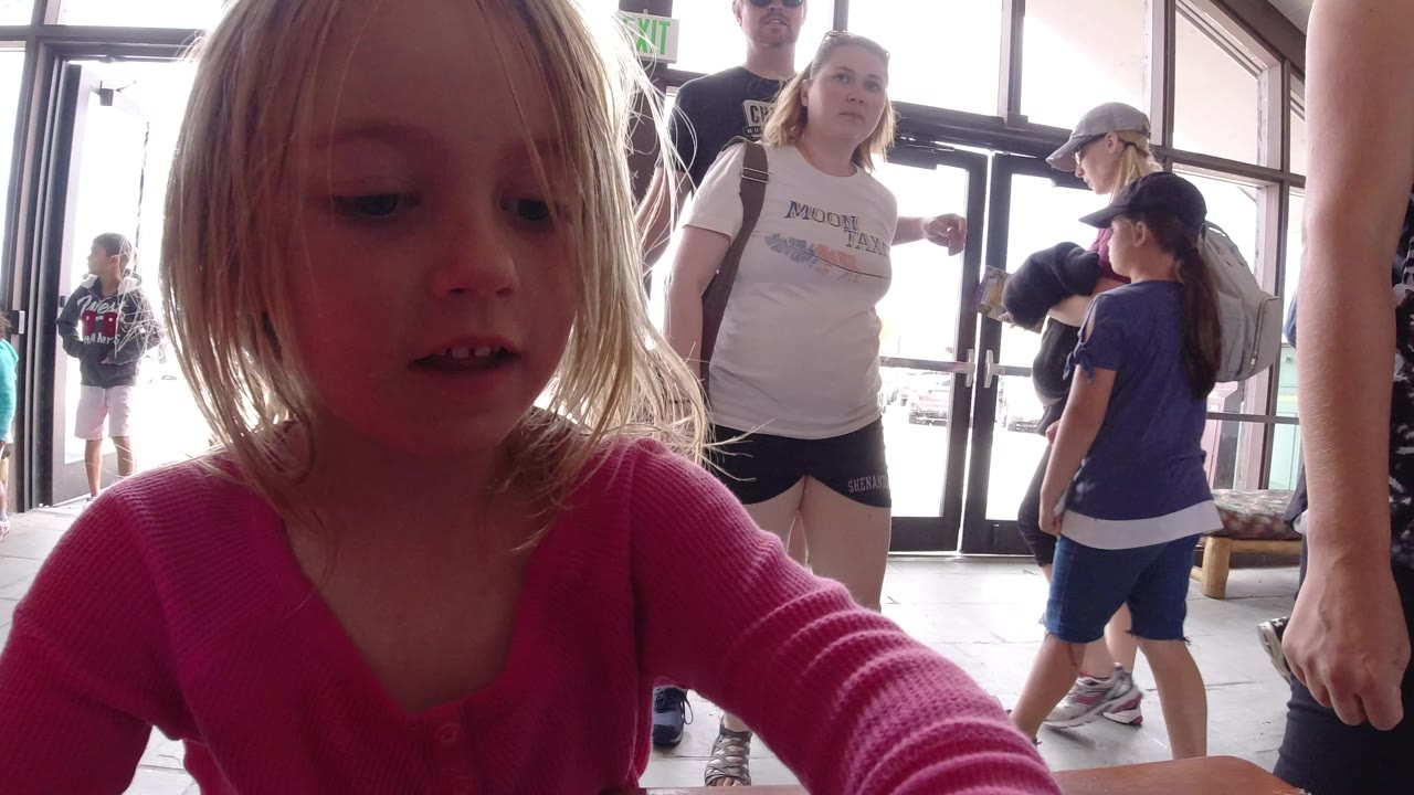 A blonde child in a pink shirt leans close to the camera at a table in Winter Park, Colorado, with adults and children standing near bright glass doors behind her.
