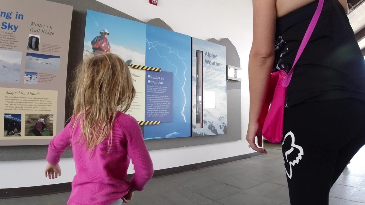 In Winter Park, Colorado, a young girl in a pink top walks toward wall displays about alpine weather while an adult stands close at frame right.