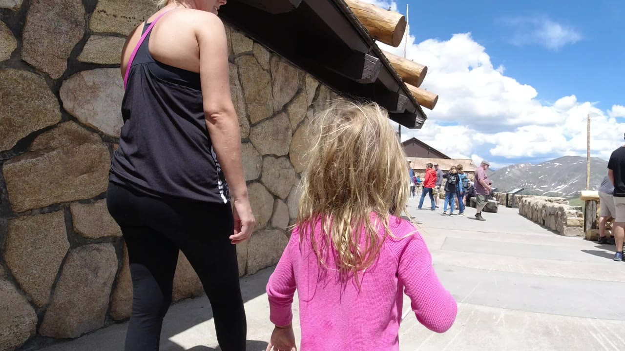 The child remains centered near the stone building as the adult companion walks at left, with a small group of tourists farther along the paved overlook area in Winter Park, Colorado.