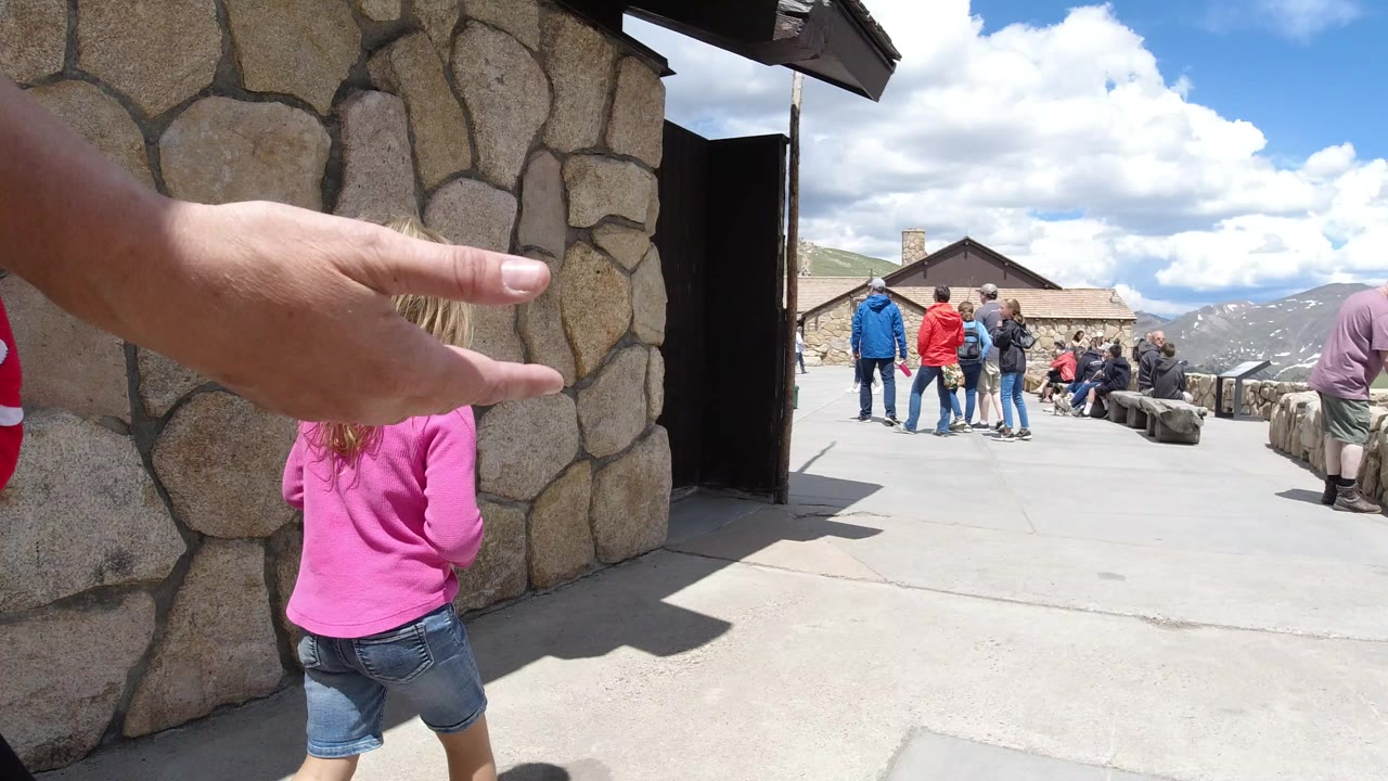 In Winter Park, Colorado, an adult hand points across the foreground while a blond child in a pink shirt stands beside a stone wall near a dark doorway, with visitors and mountains beyond.