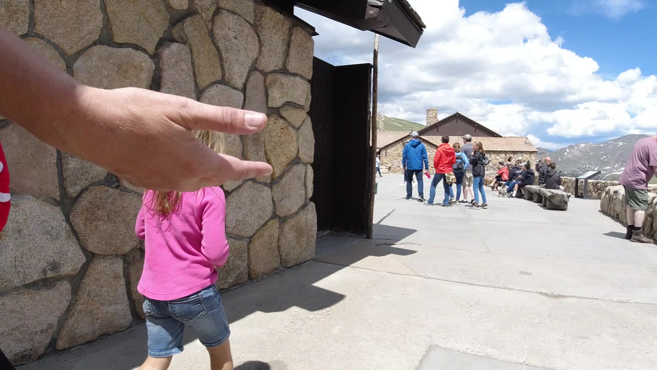 The adult hand stays extended in the foreground as the child faces the open deck, with a stone entrance at left and groups of visitors spread across the Winter Park overlook.
