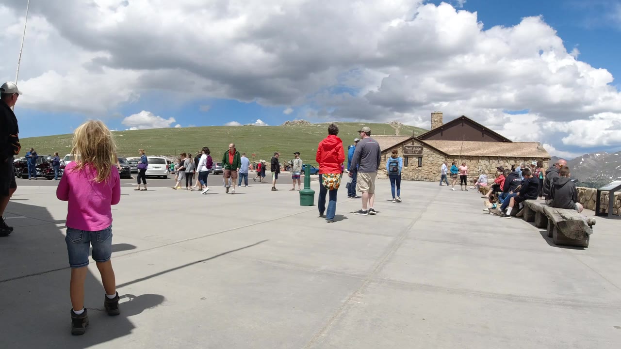 Young girl in pink standing on pavement at an outdoor event with people and hills behind her.