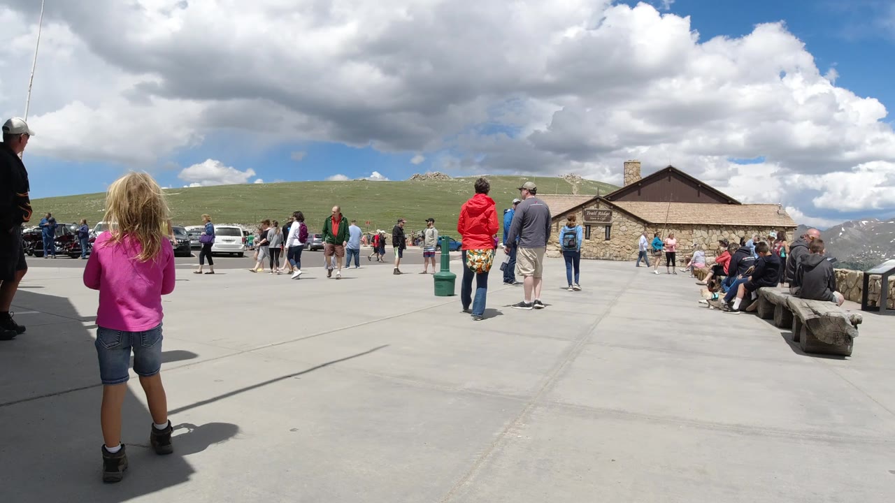 The girl is seen from behind near the center foreground in Winter Park, Colorado, facing a loose crowd, parked vehicles, a stone building, green hills, and bright cloud cover.