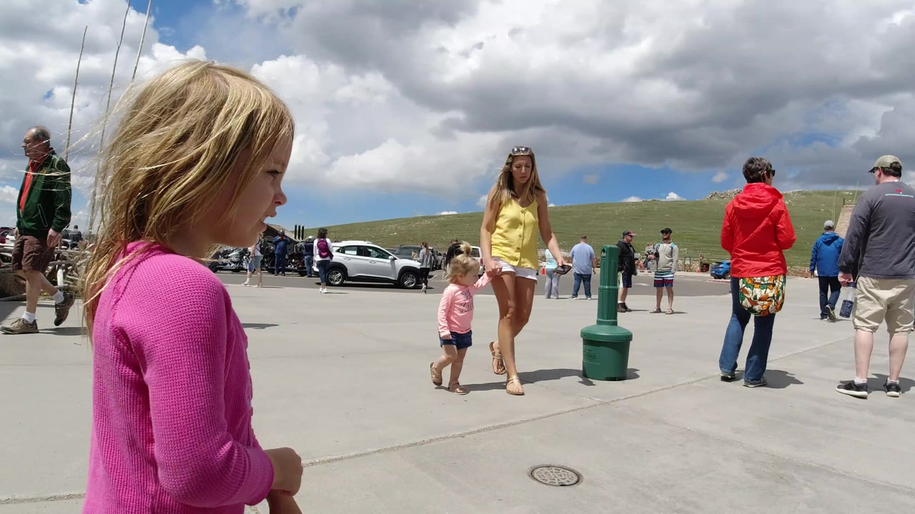 A blonde child in a pink long-sleeve top stands in the left foreground of a Winter Park, Colorado plaza while adults and children gather near cars under large clouds.