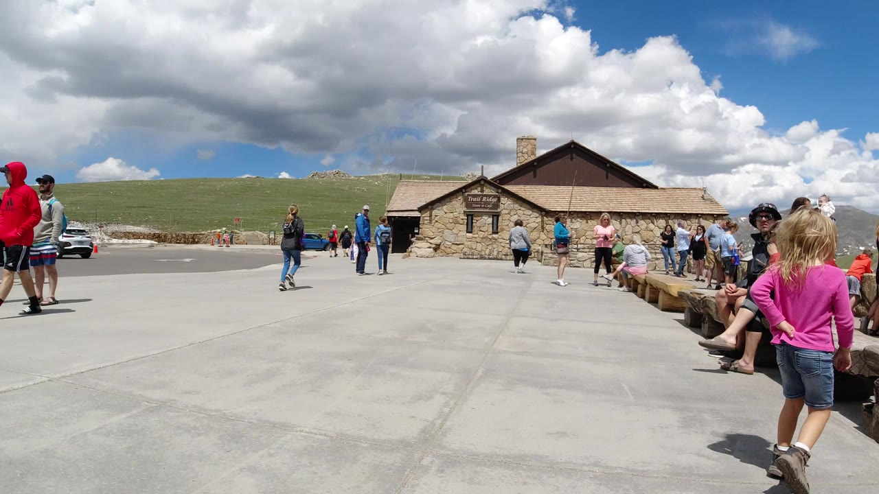 Winter Park, Colorado visitors stand and sit outside a stone building with benches, open pavement, grassy hills, and a bright cloud-filled sky.