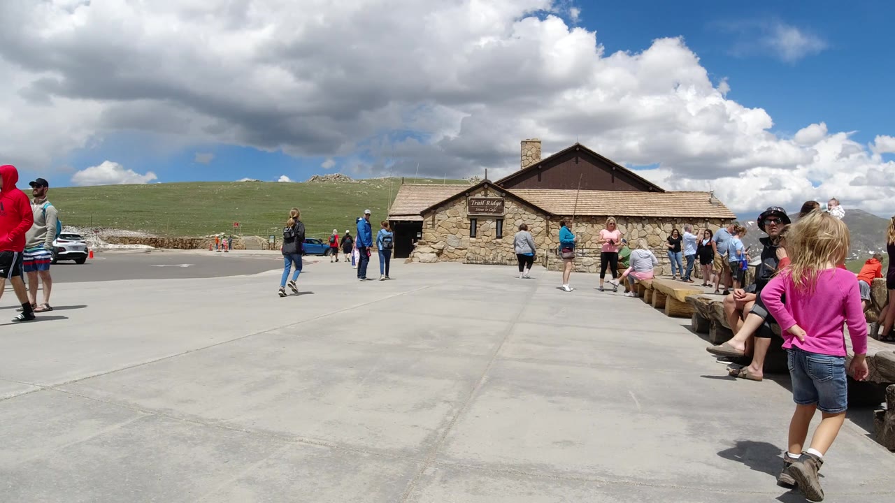 Winter Park, Colorado overlook scene with visitors around a stone building, a child in a pink top in the foreground, and mountains beneath broken clouds.