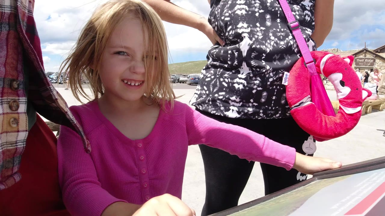 The young girl looks toward the camera while holding near the sign edge, with a bright pink bag, cropped adults, parked visitors, and sky visible in Winter Park, Colorado.