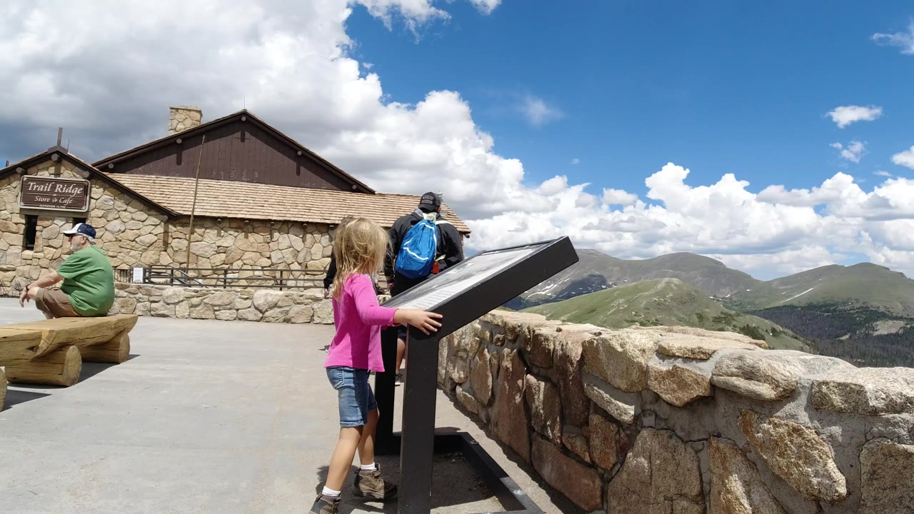 The girl remains beside the angled sign as an adult with a blue backpack faces the mountain view and a seated adult stays near the stone building.