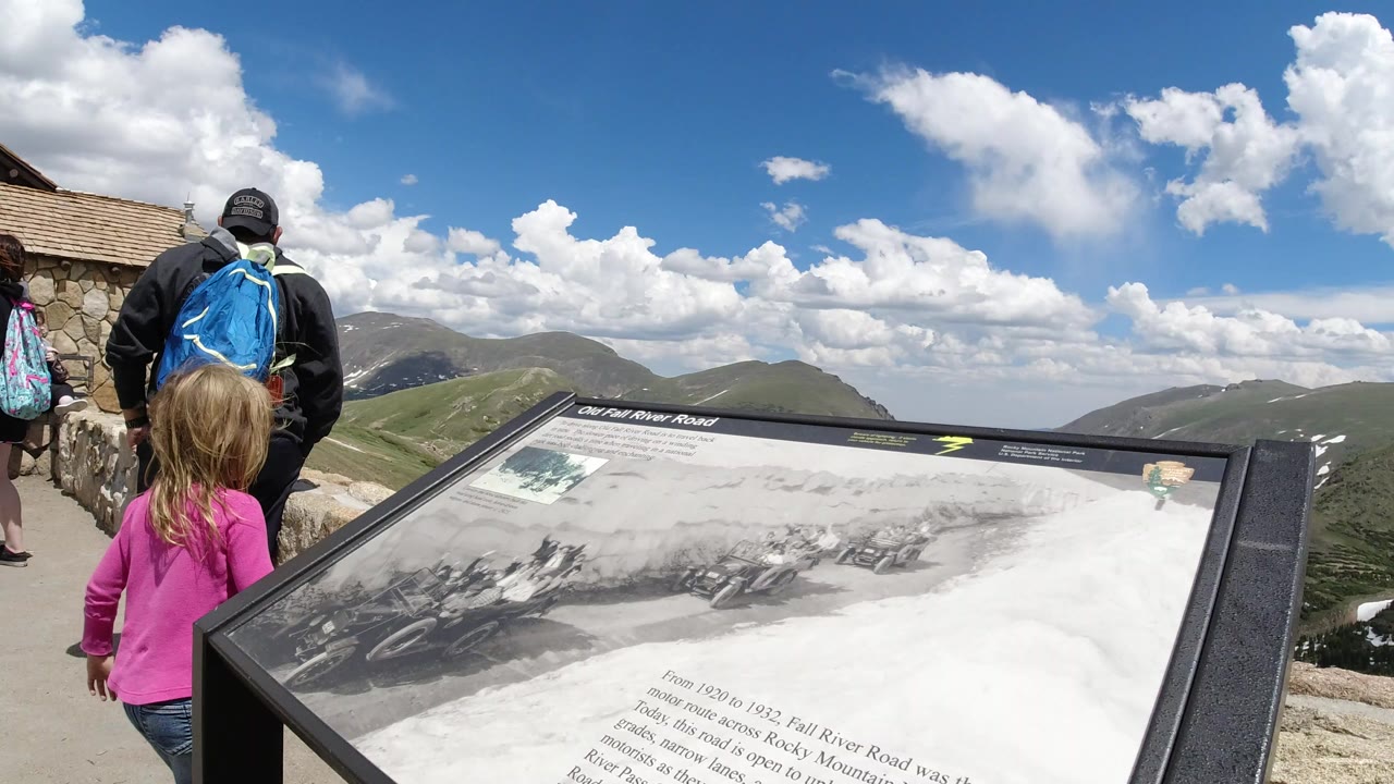 At 0:00 in Winter Park, Colorado, a child in pink and an adult with a blue backpack stand near a stone overlook wall beside an Old Fall River Road interpretive sign, with mountain ridges and clouds beyond.