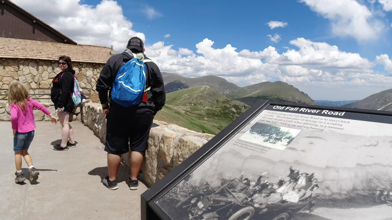 At Winter Park, Colorado, visitors stand near a stone wall and an Old Fall River Road interpretive sign while mountains and clouds fill the background.