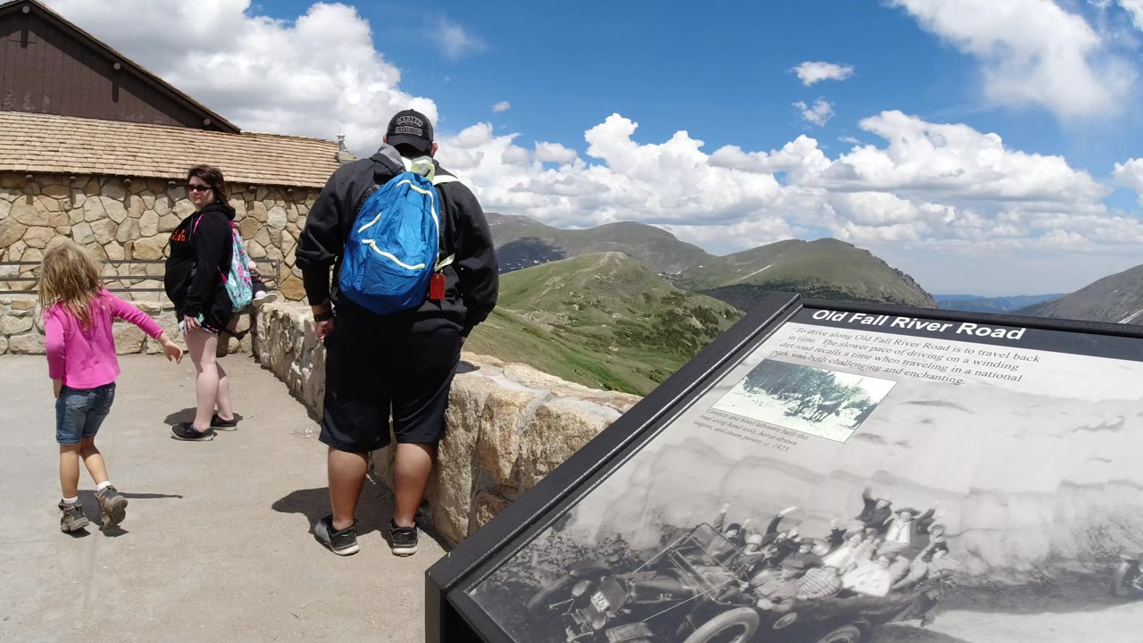 A child in a pink top and adults walk along a stone overlook wall in Winter Park, Colorado, with green ridges and bright clouds beyond the walkway.