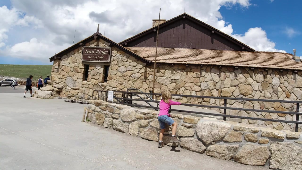 A young girl in a pink shirt stands at a low stone wall outside a stone building in Winter Park, Colorado, with the Trail Ridge Store and Cafe sign visible and a few people in the distant left background.