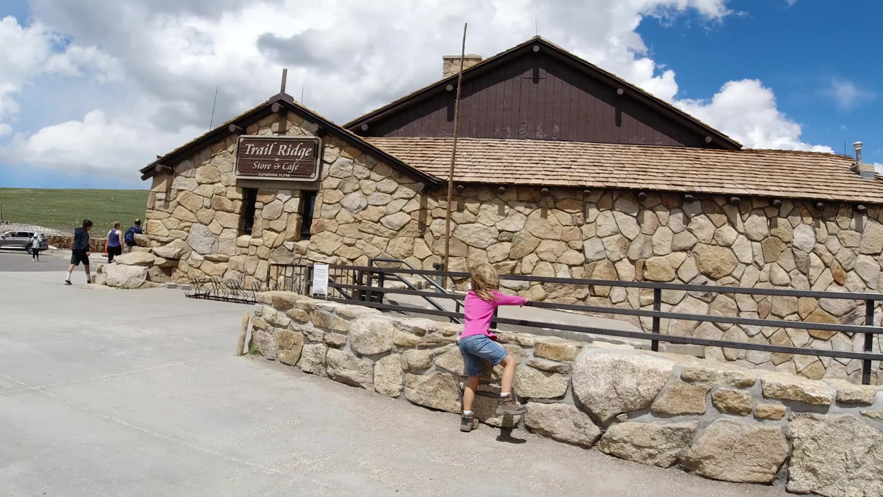The girl faces the stone wall in Winter Park, Colorado, with one leg lifted as she prepares to climb, while the stone storefront, rail fence, blue sky, and clouds fill the background.