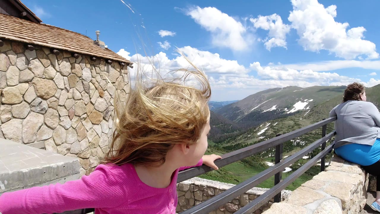 Blond child in pink standing by a deck railing above mountains.