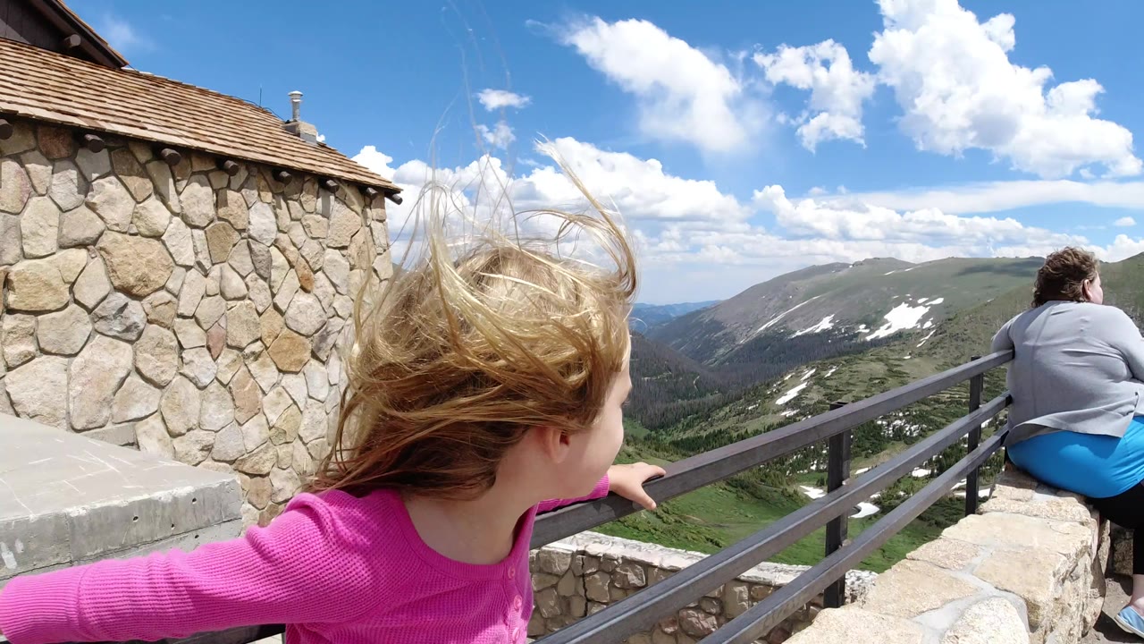 At 0:01, the child remains near the railing with windblown hair as the stone wall, seated adult, distant ridges, snow patches, and bright clouds stay in view.