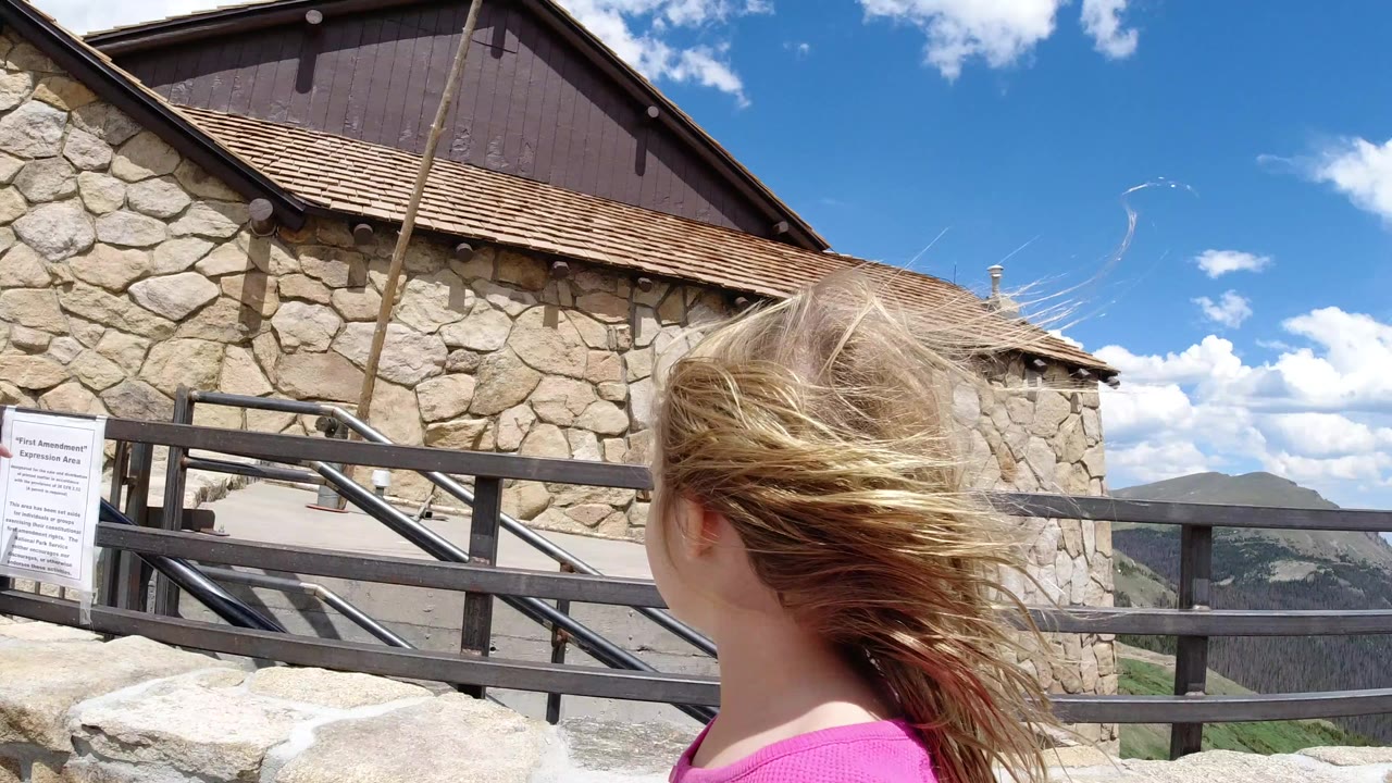 Blonde child beside a stone building and metal railing under blue sky.