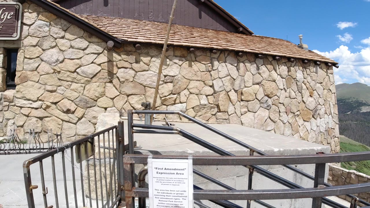 Winter Park, Colorado exterior view of a tan stone wall, wood roofline, metal ramp railings, foreground sign, distant hills, and no visible people.