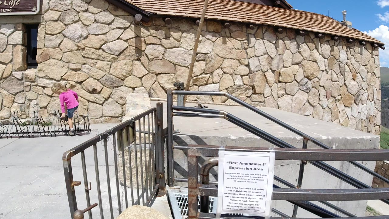 A child in a pink top bends over a black bicycle rack on a concrete walkway in Winter Park, Colorado, with a stone wall, metal railings, and a posted sign in the foreground.