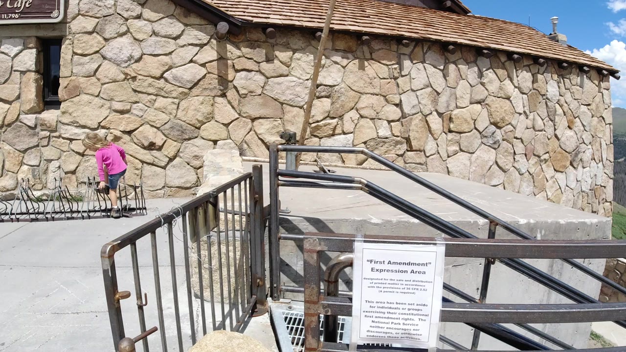 The child remains near the bicycle rack in Winter Park, Colorado, with the stone facade filling the background and the foreground sign and railings still visible.