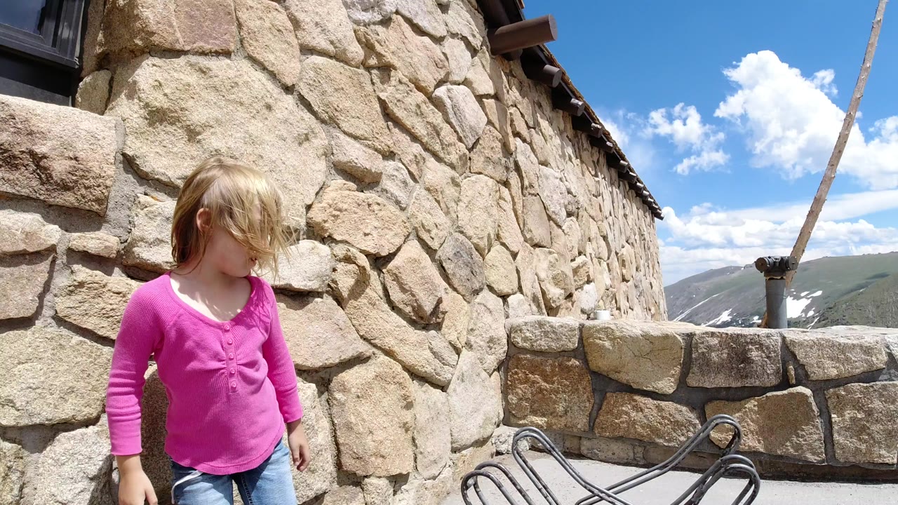 A young blond girl in a pink shirt stands near a rough stone building wall in Winter Park, Colorado, with blue sky and distant mountains beyond a low stone barrier.