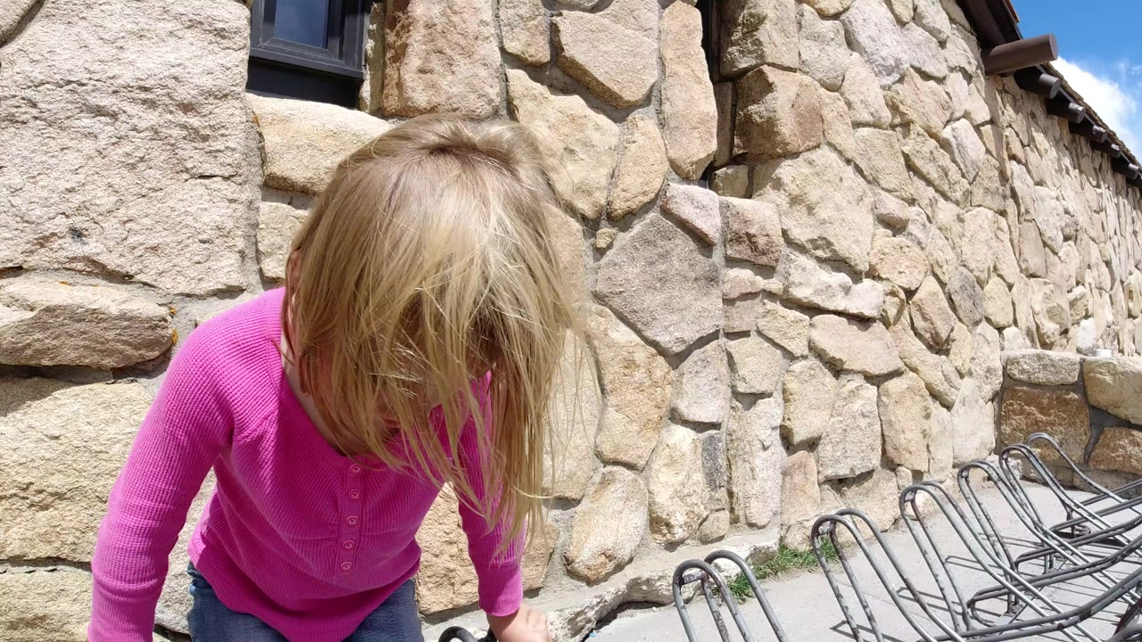 In Winter Park, Colorado, a blonde child in a pink top leans forward near rows of metal chairs against a rough stone wall in bright daylight.