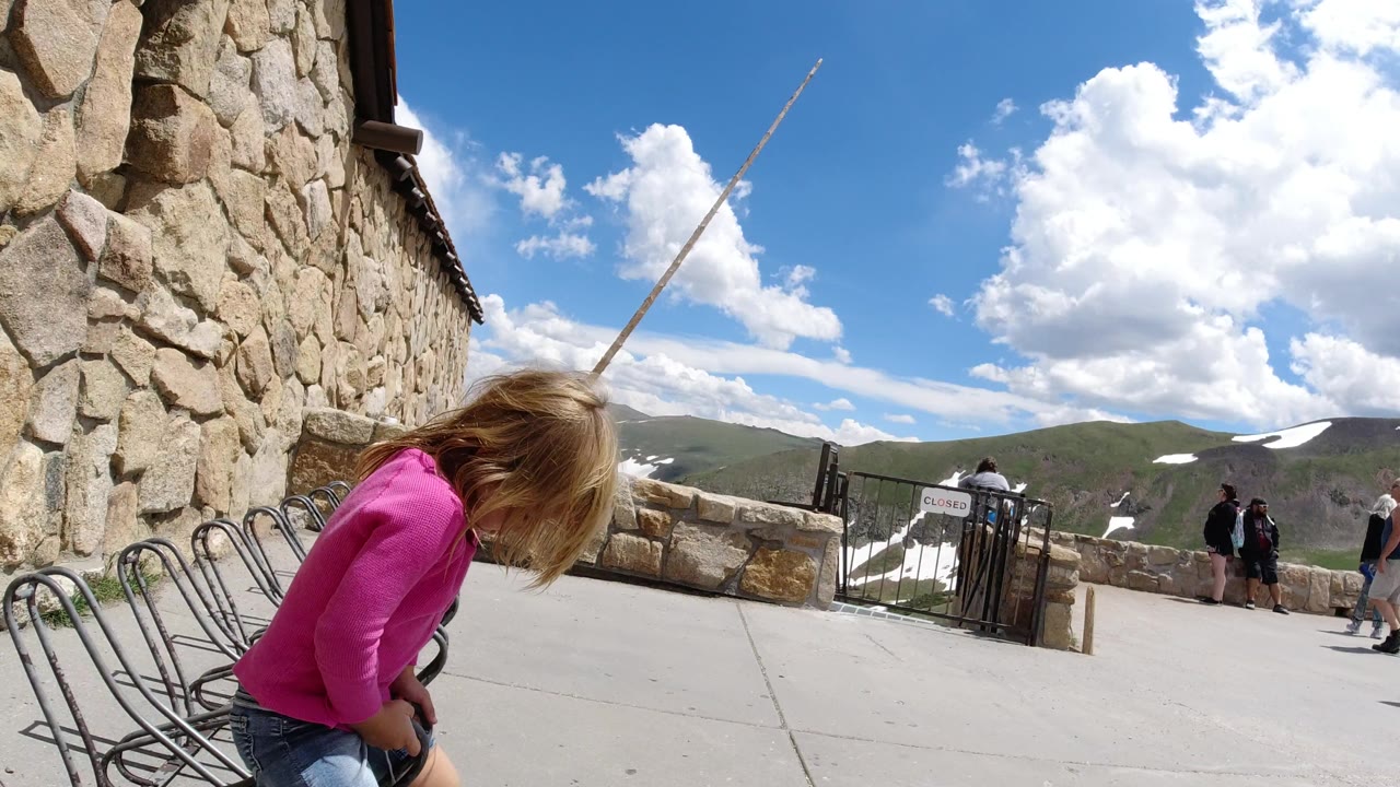 At Winter Park, Colorado, the child leans farther forward beside the bike racks, while the stone building wall, paved overlook, distant mountains, snow patches, and a few visitors remain in view.