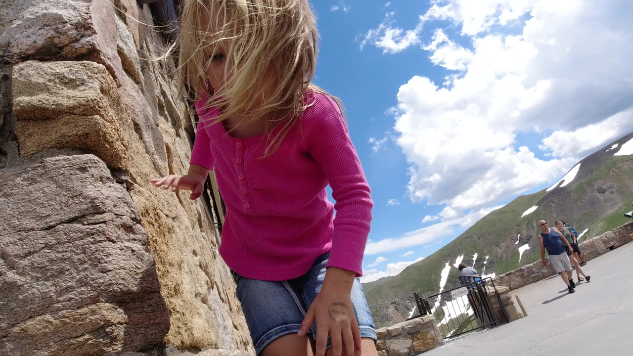 Blonde girl in pink climbing beside a stone wall under a bright sky.
