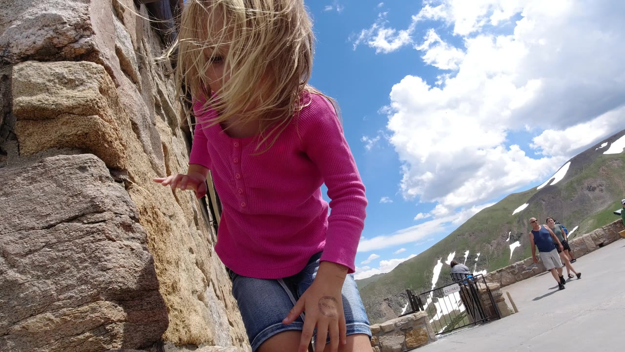 At 0:01 in Winter Park, Colorado, the girl keeps one hand near the stone wall while her hair covers part of her face and several visitors walk along the open paved area behind her.