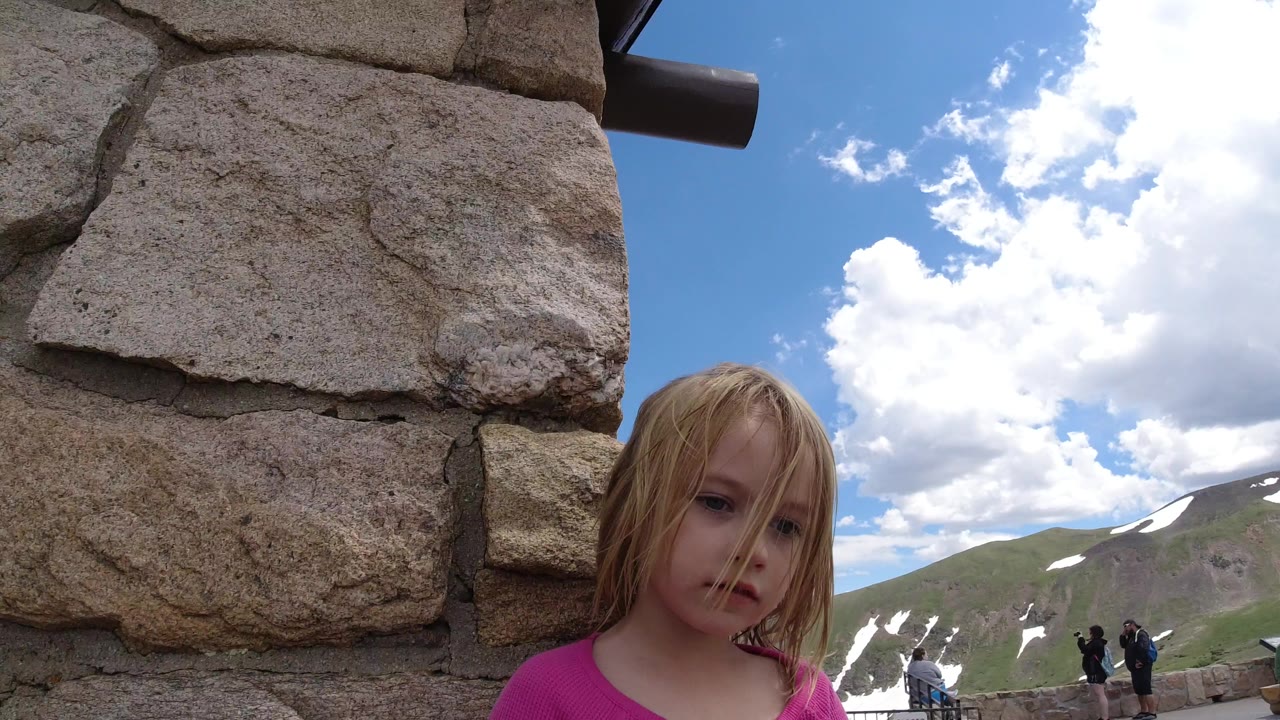 Young girl beside a stone wall with mountains and clouds behind her.