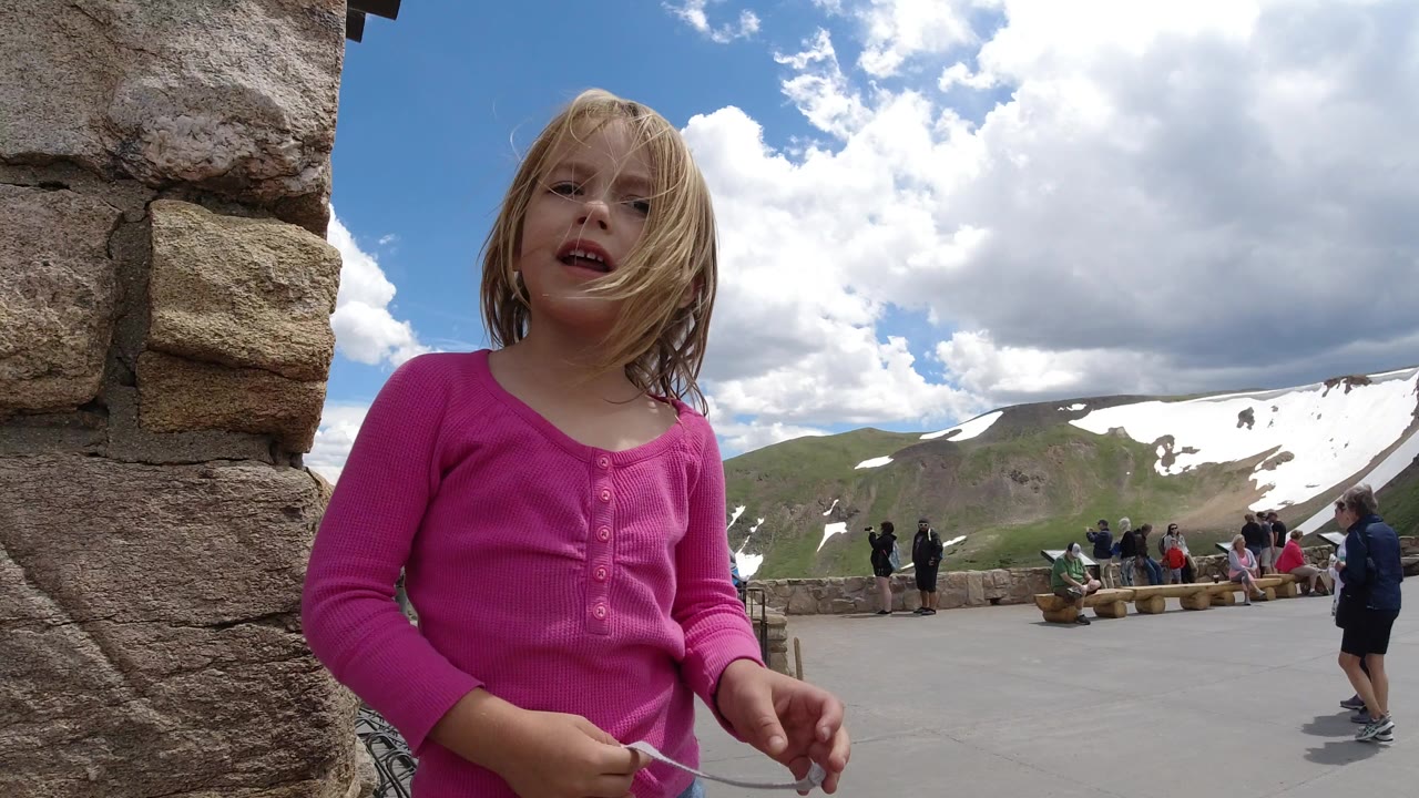 Young girl in pink standing by a stone wall with mountains and clouds behind her.