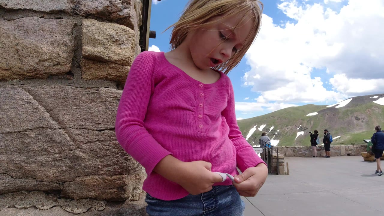 The girl keeps her gaze down and handles a small object beside the rough stone wall, while distant visitors stand at the Winter Park, Colorado mountain viewpoint.