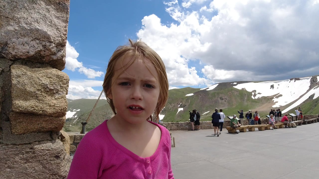 Young girl in a pink shirt standing beside a stone wall at a mountain overlook.