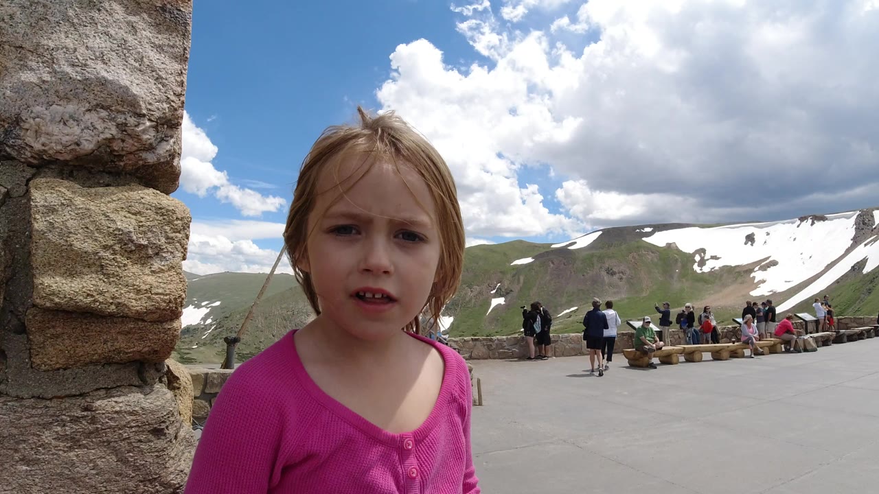The girl remains close to camera near the stone wall in Winter Park, Colorado, with her face centered and the overlook crowd and snowy mountain ridge spread behind her.