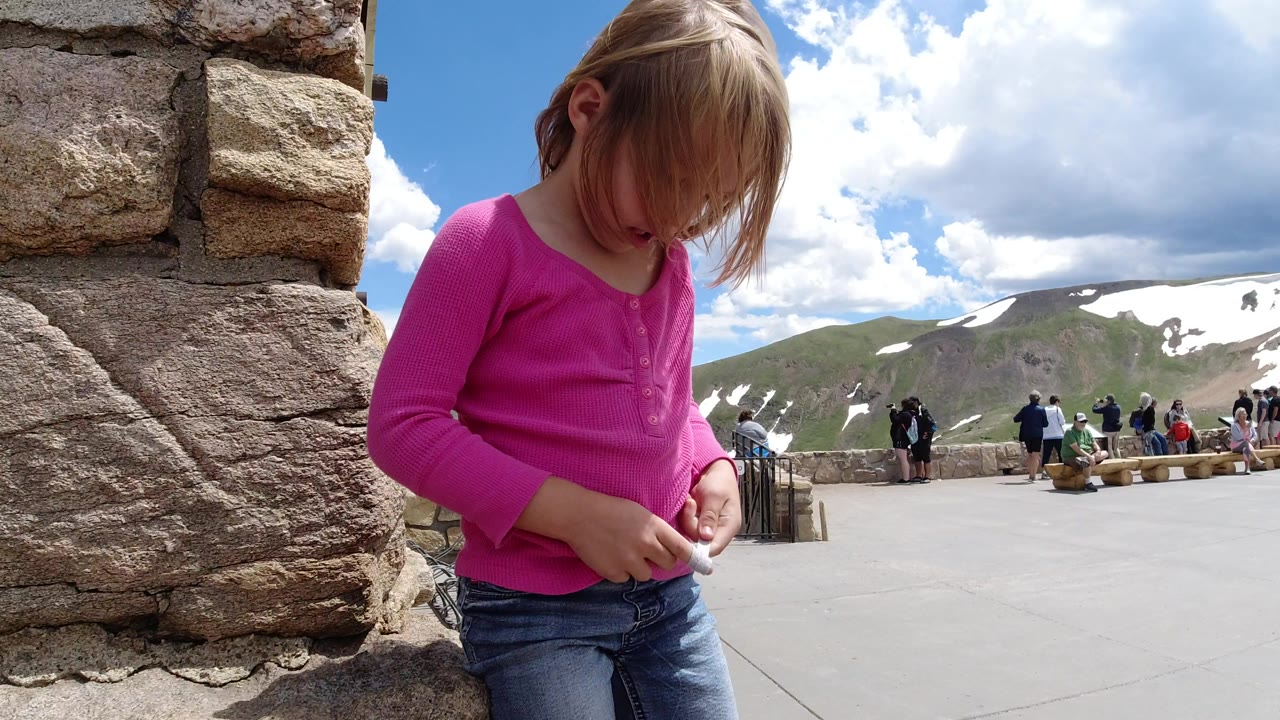 At a Winter Park, Colorado overlook, a young girl in a bright pink shirt looks down while holding string beside a rough stone wall, with snowy mountains and visitors behind her.