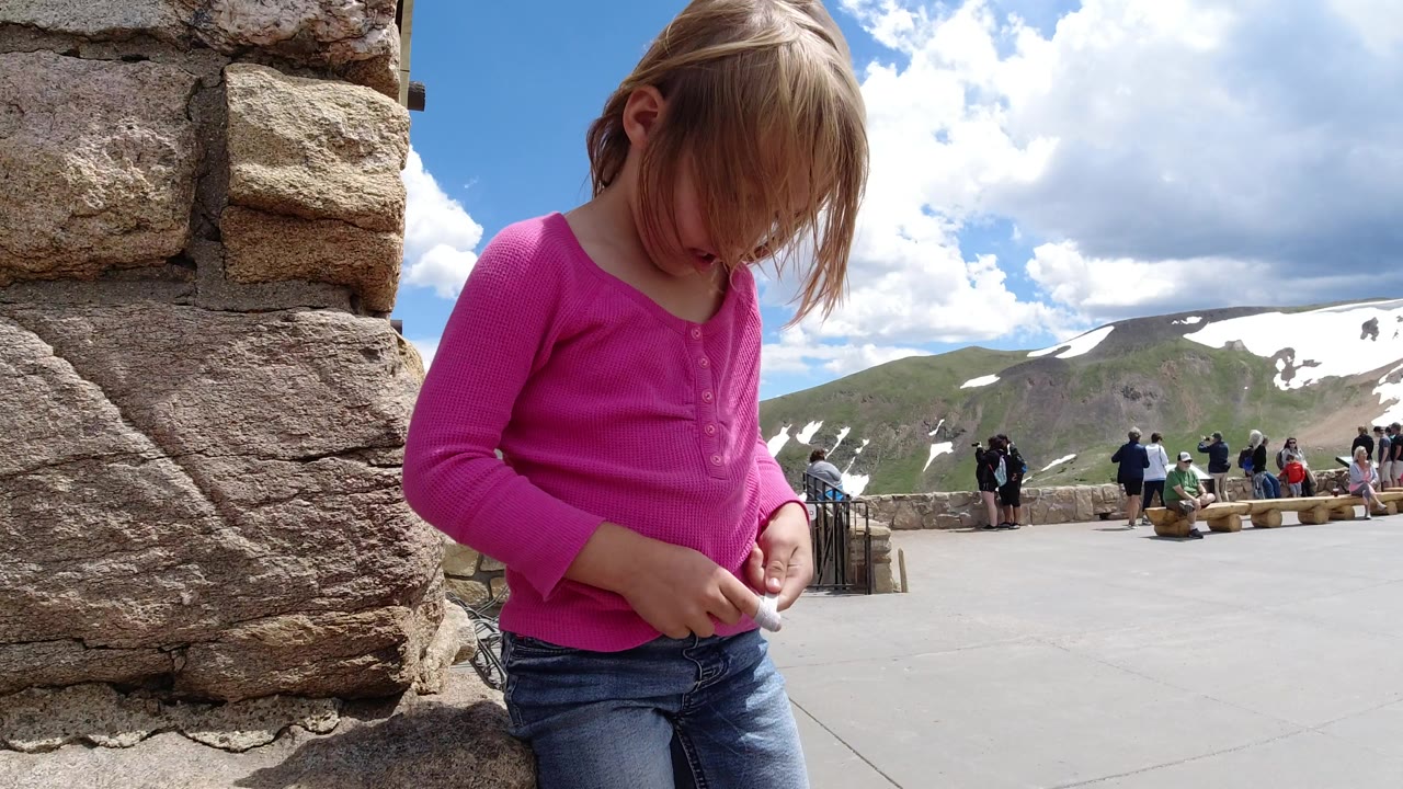 The girl remains near the stone wall in Winter Park, Colorado, looking down at the string as the paved overlook opens toward snow-streaked mountains and small visitor groups.