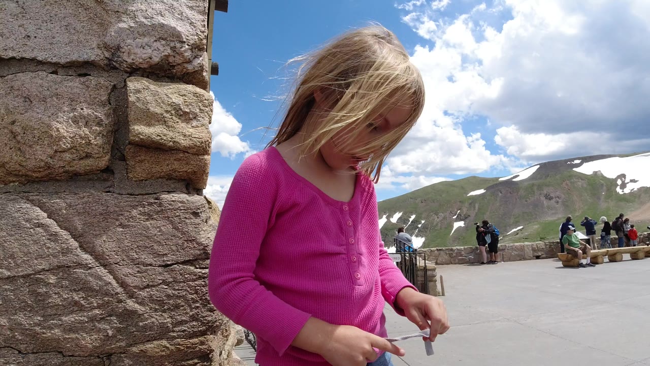 Young girl in a pink shirt holding rope beside a stone wall at a mountain overlook.