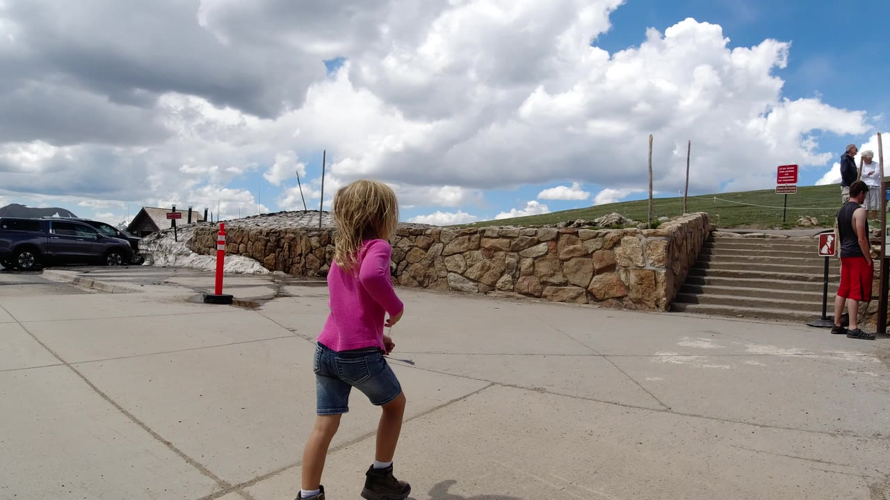 The girl in pink continues toward the stone steps in Winter Park, Colorado, as adult visitors stand near a display board beside the rock wall.
