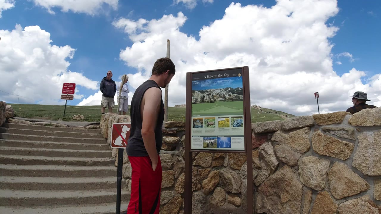 Young male in red shorts stands near a brown information sign by stone stairs in Winter Park, Colorado, with two adults visible at the top of the steps.