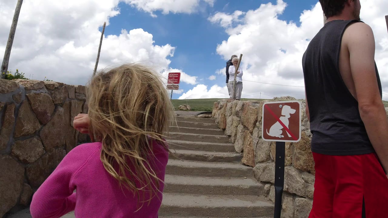A young girl in pink moves toward the stairs in Winter Park, Colorado as two older adults stand higher up near a rope line and red posted signs.