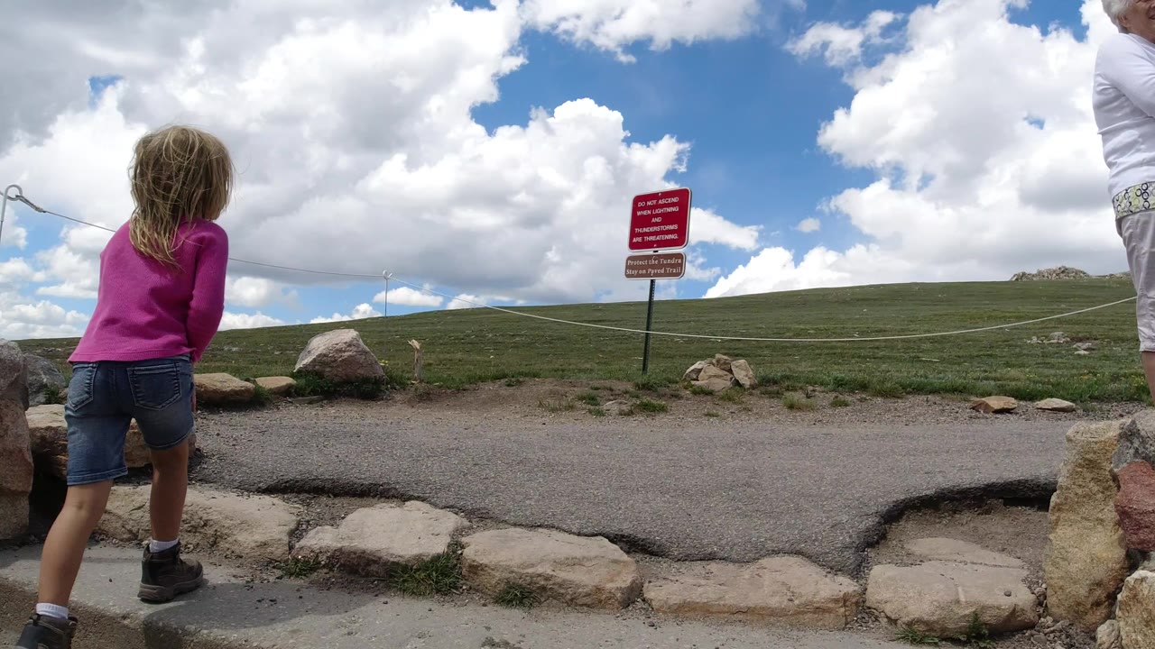 Blonde girl facing a chain barrier beside rocks and a red warning sign.