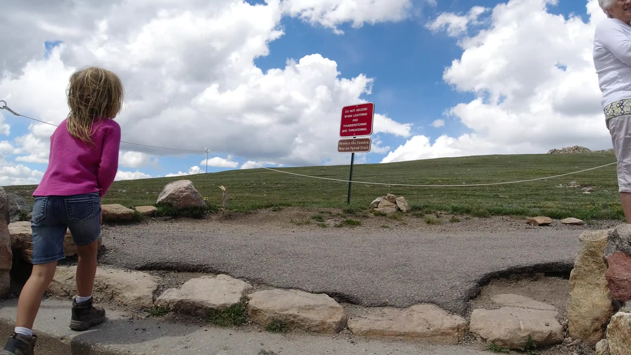 The girl stands near the low chain in Winter Park, Colorado as a partly cropped adult passes near the top edge, with rocks, grass, and a red warning sign ahead.