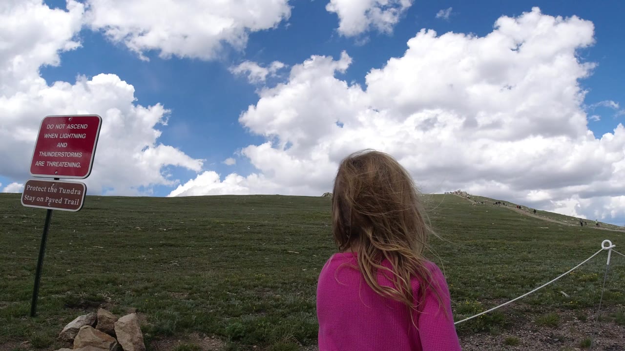 Young girl in a pink top walking toward a grassy hillside trail sign.