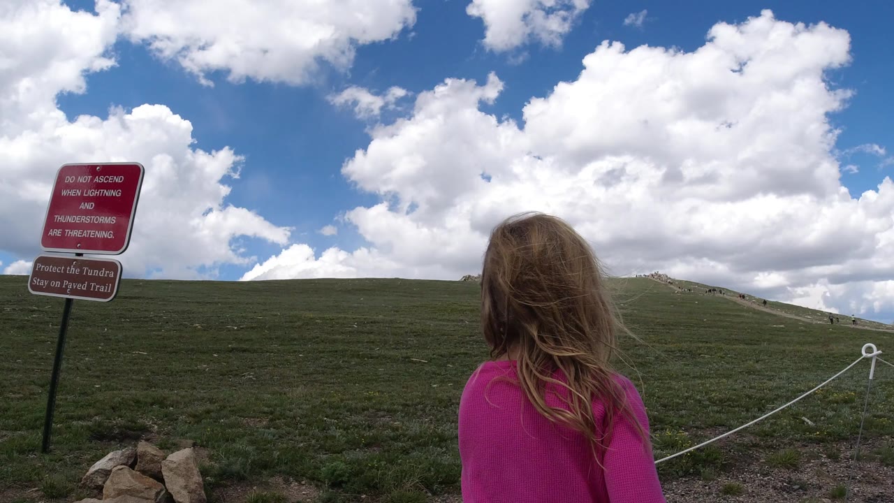 The young girl remains in the foreground while the Winter Park hillside stretches ahead, with a red trail warning sign at left and a rope boundary crossing the grass.