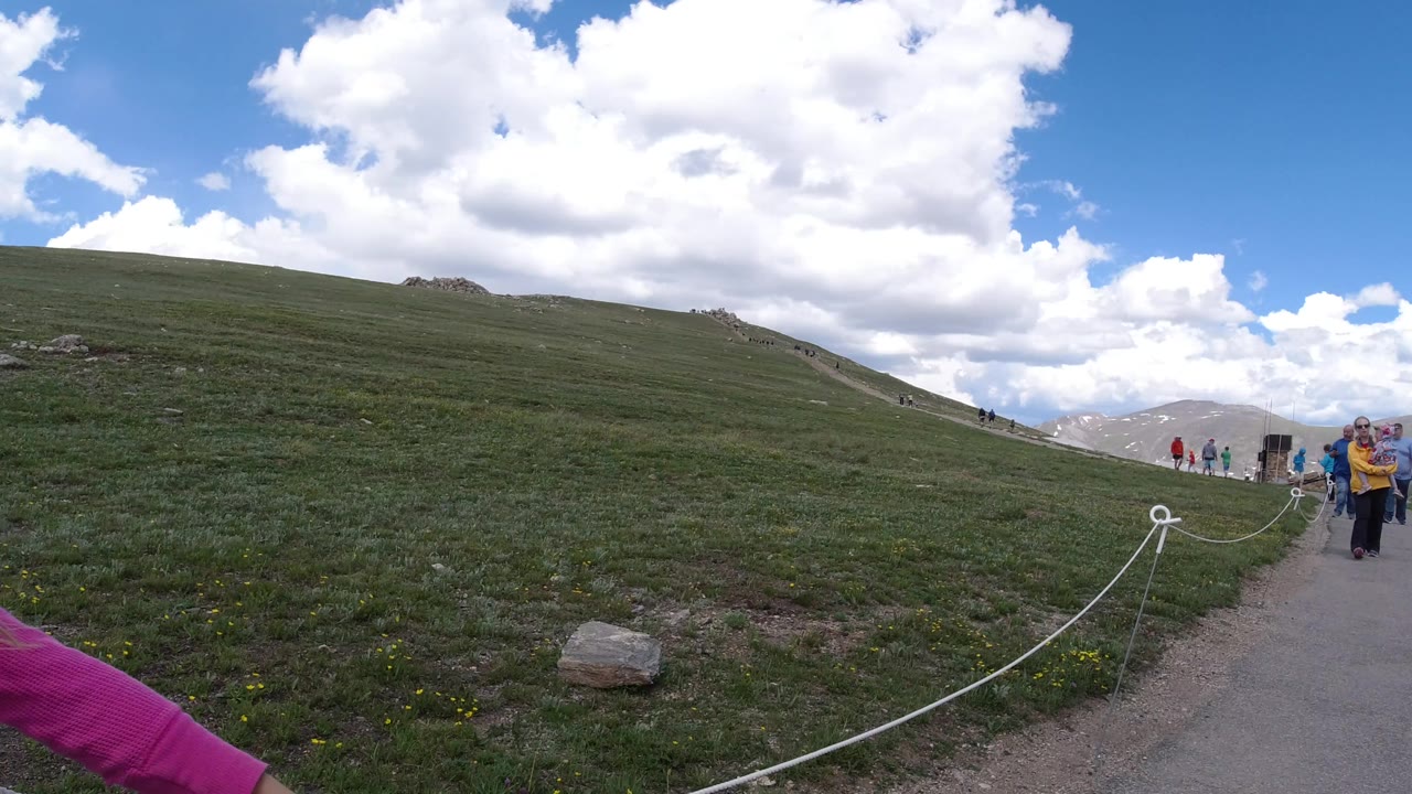 A wide Winter Park, Colorado path scene shows a white rope barrier leading toward distant visitors, with a low grassy slope under a blue sky filled with large clouds.