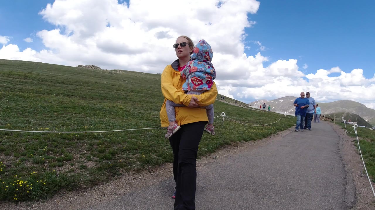 At the start of the Winter Park, Colorado clip, a woman carries a child beside a paved path with grassy hillside, rope barrier, distant walkers, and bright clouds.