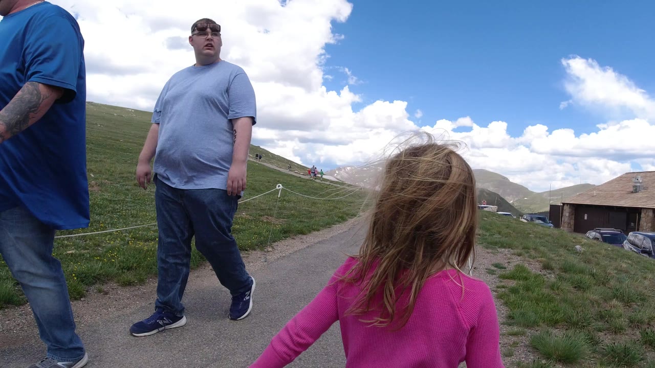The girl in pink remains in the foreground on a Winter Park path as an adult man in gray walks past and another adult in blue is partly cropped at the edge.