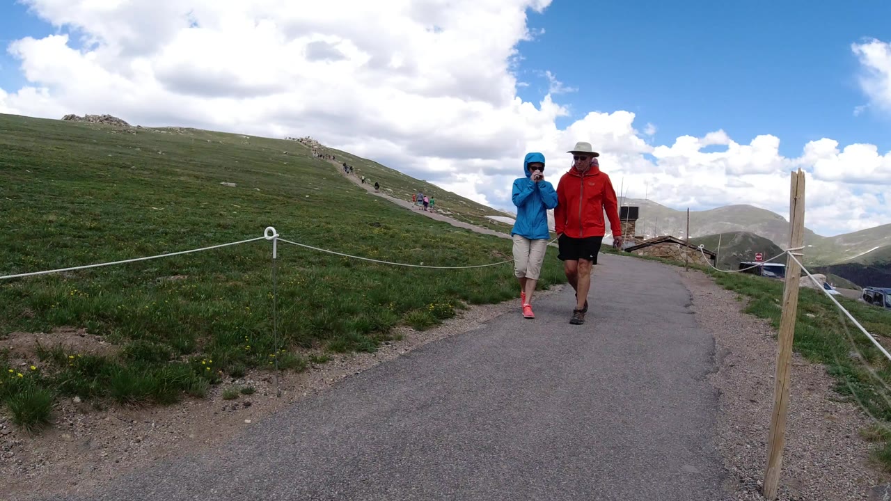 An adult in a red jacket and a child in a blue jacket walk toward the camera on a paved trail in Winter Park, Colorado, with rope fencing and grassy slopes around them.