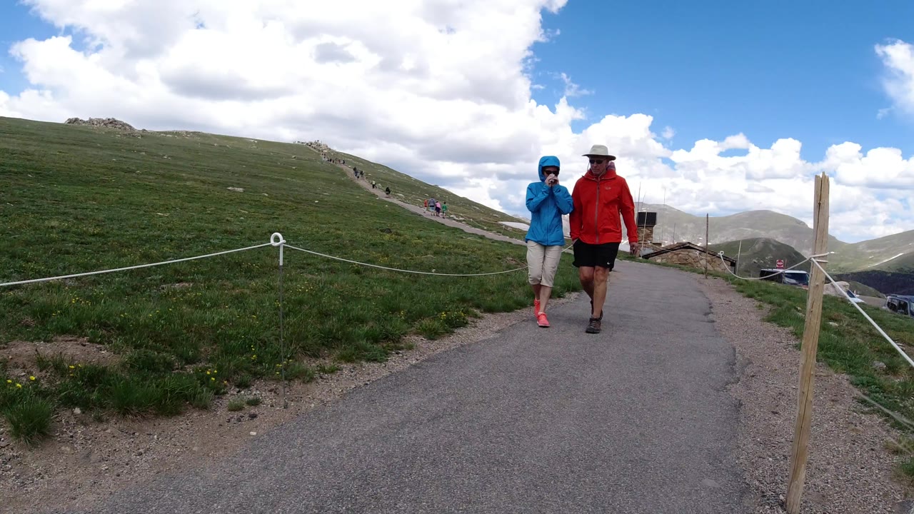 The adult and child remain near the right side of the paved Winter Park trail, with a sloping green hillside, rope barriers, and bright clouds filling the scene.