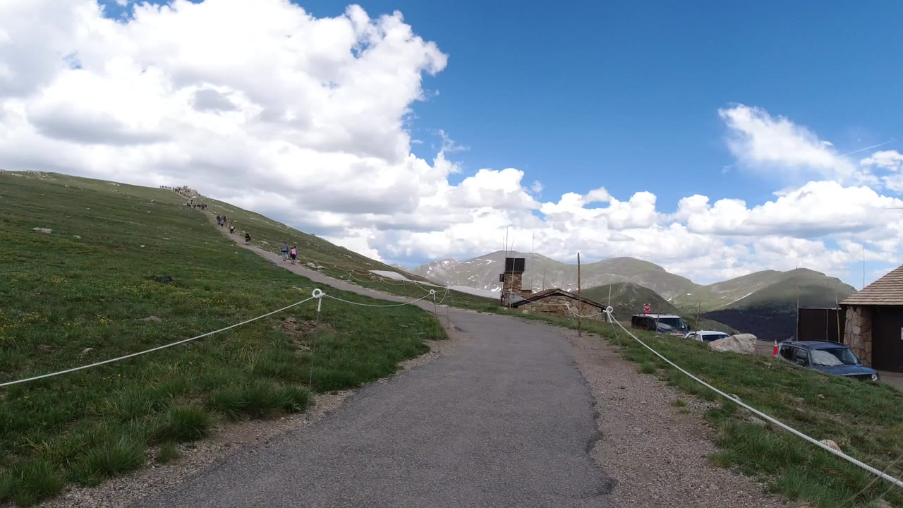 Paved mountain road beside grassy slope under cloudy blue sky.